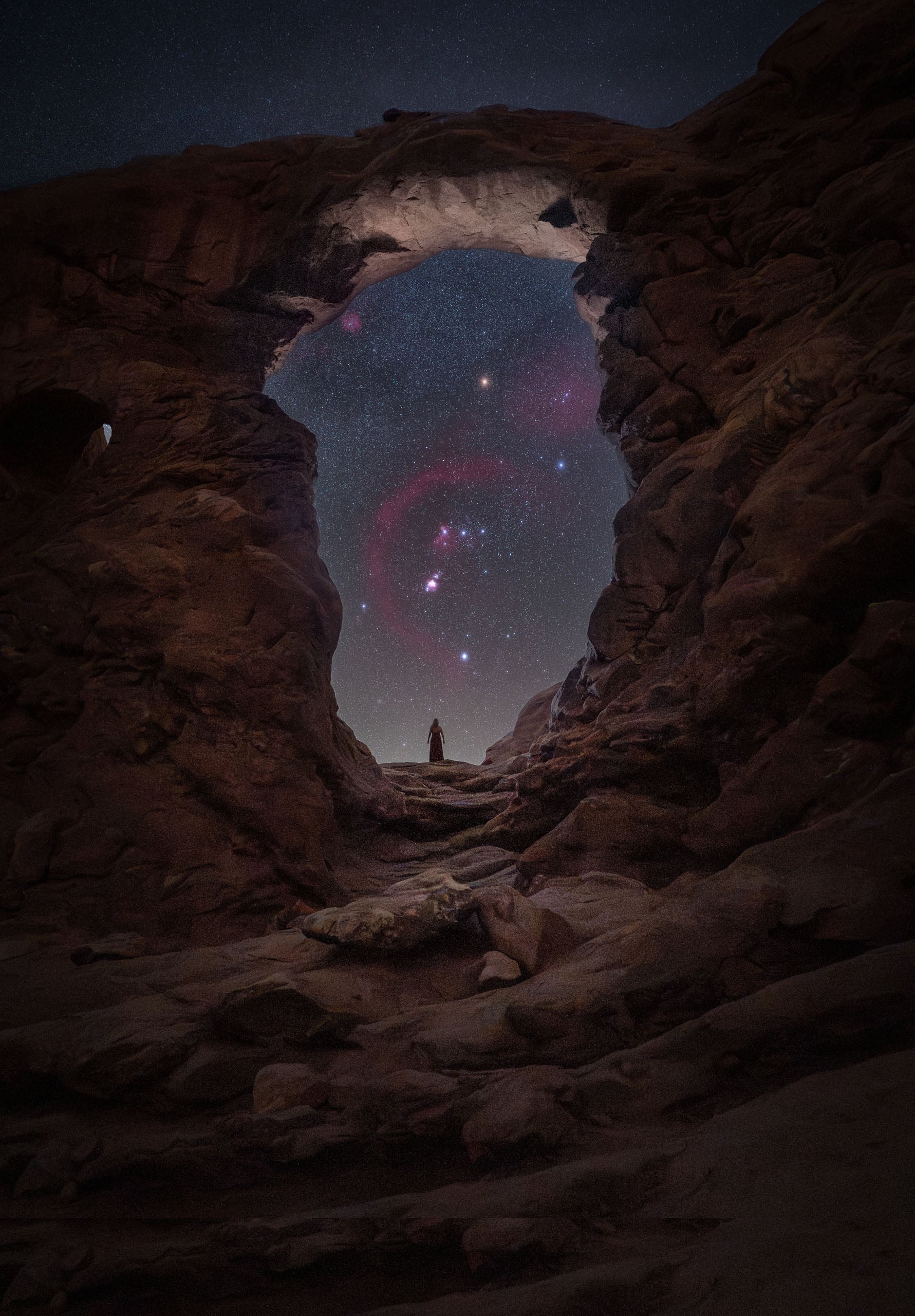 A person stands in the distance beneath a large natural rock arch, looking out at a night sky filled with stars and the Orion constellation.