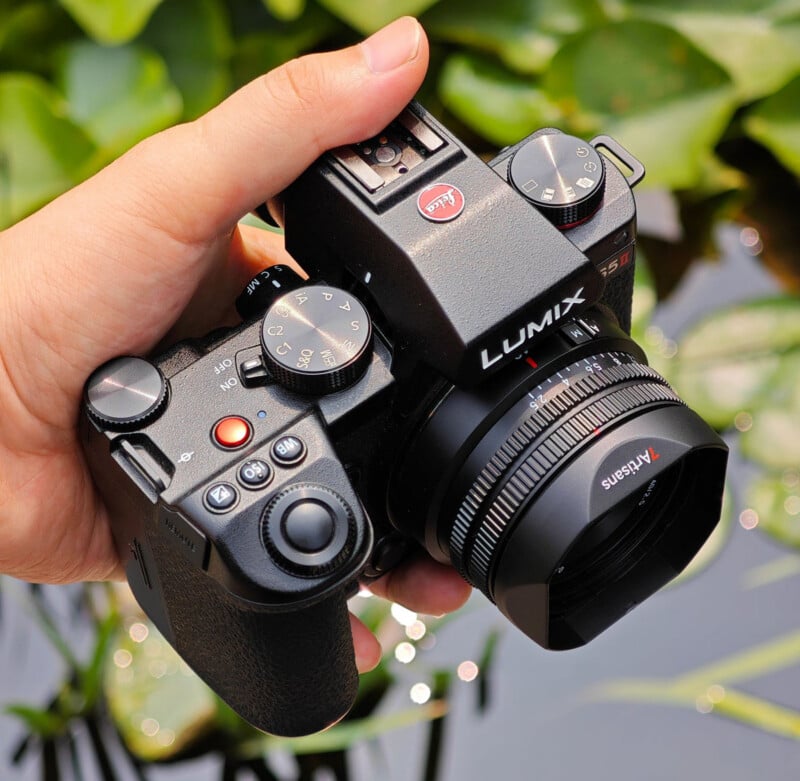 A hand holds a black Lumix digital camera with a 7Artisans lens attached, set against a blurred background of green foliage.