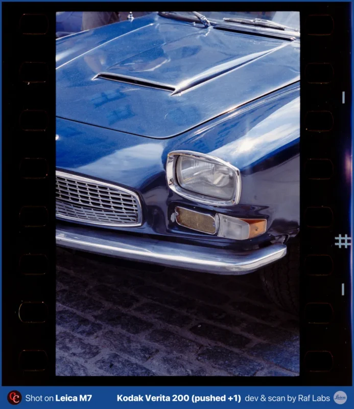 A close-up view of the front corner of a blue vintage car parked on a cobblestone street, framed by film sprocket holes.