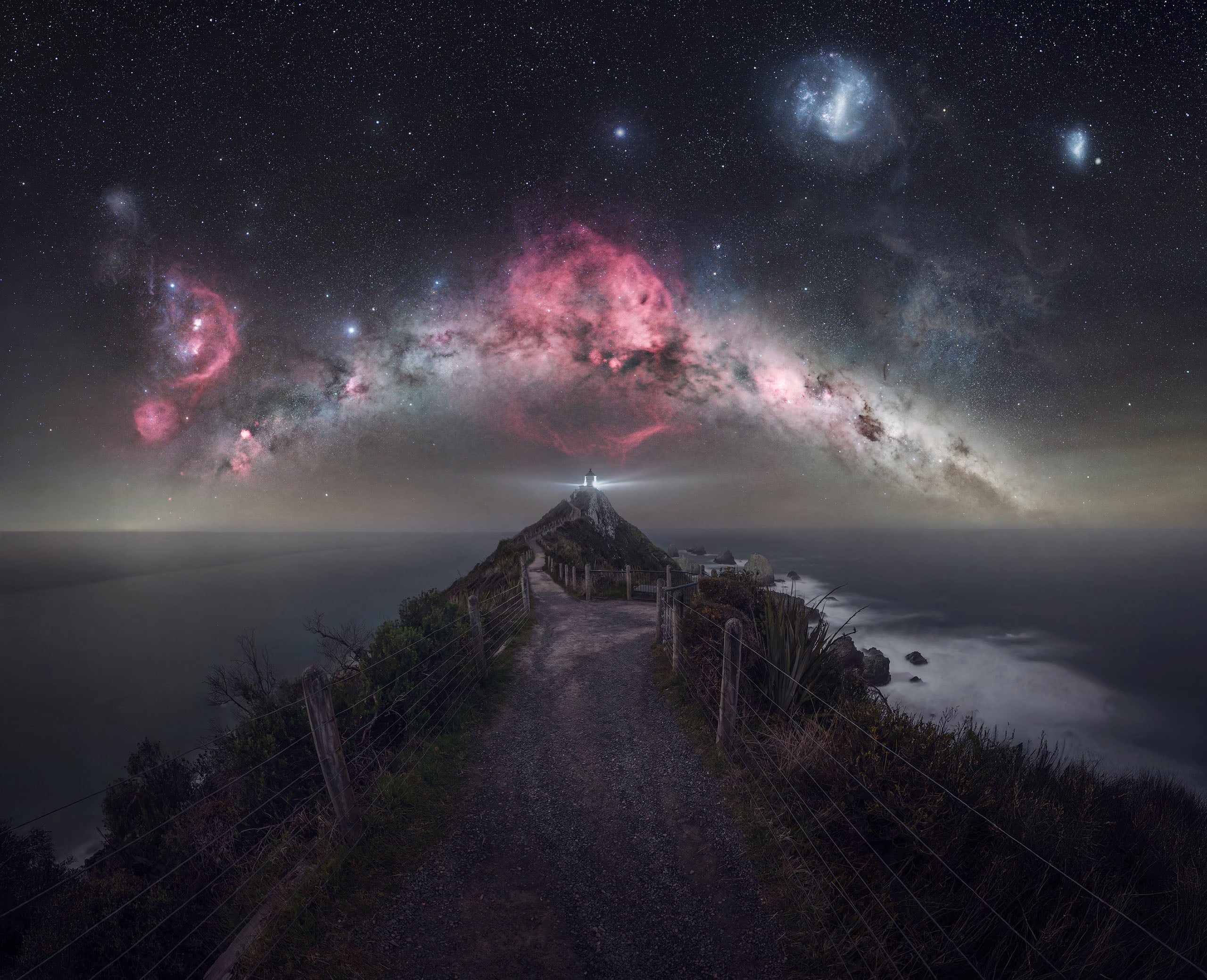 A lighthouse sits at the end of a narrow, fenced path overlooking the ocean beneath a vibrant, star-filled night sky.