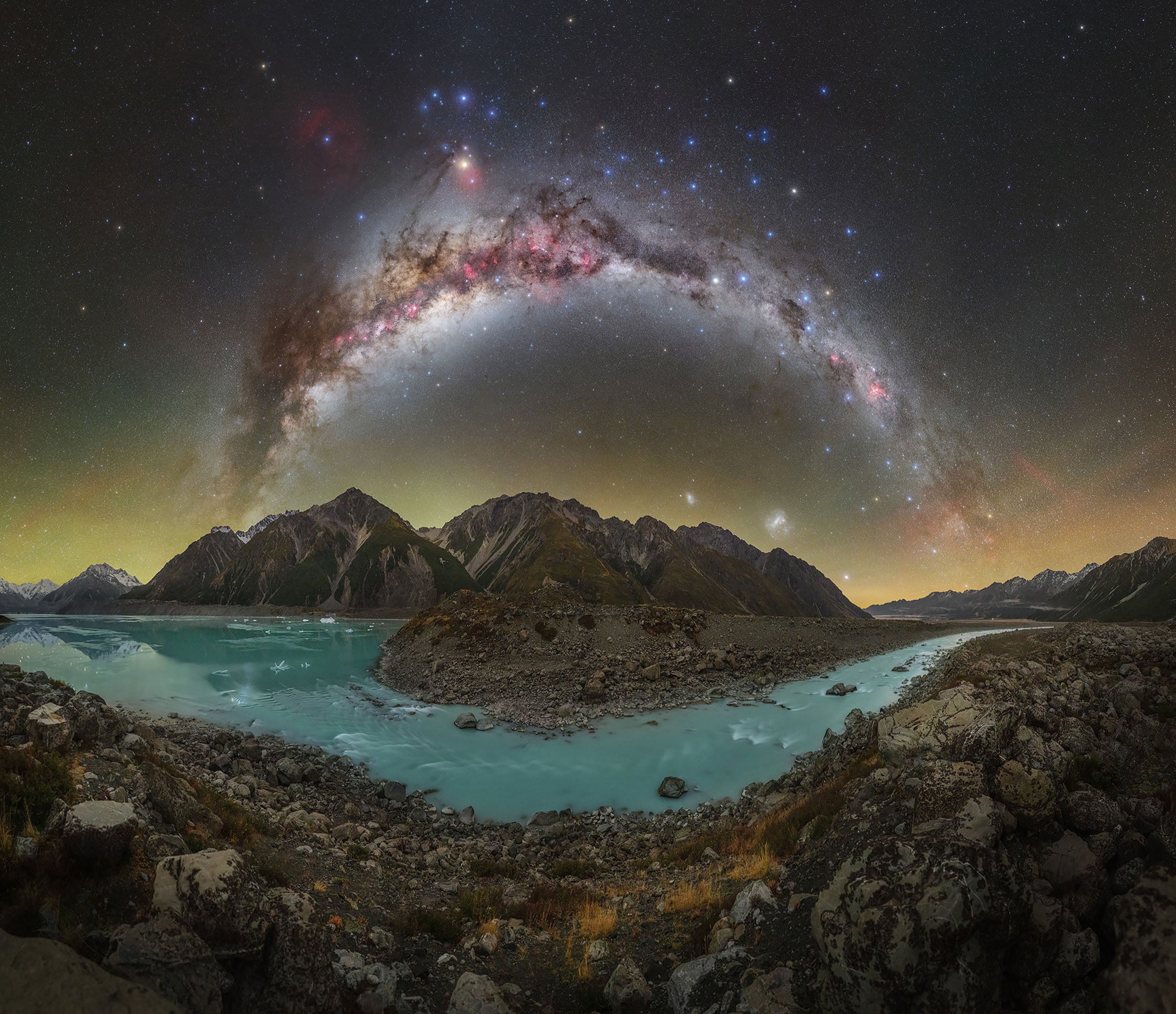 A panoramic view of a turquoise glacial lake and rocky shoreline beneath a vibrant, arched Milky Way galaxy over mountains.