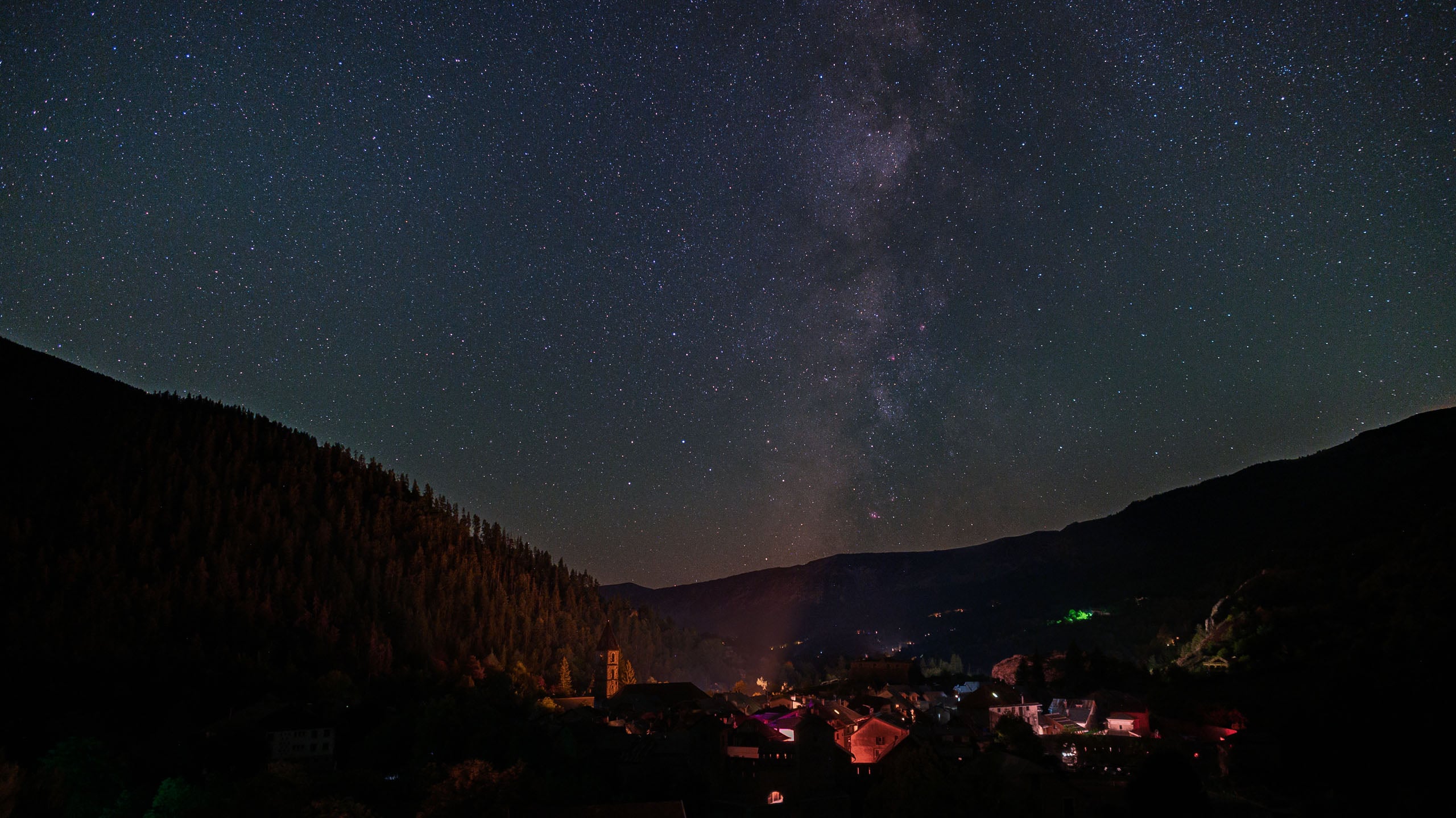 A small village nestled in a dark mountain valley under a clear night sky filled with stars and the Milky Way.