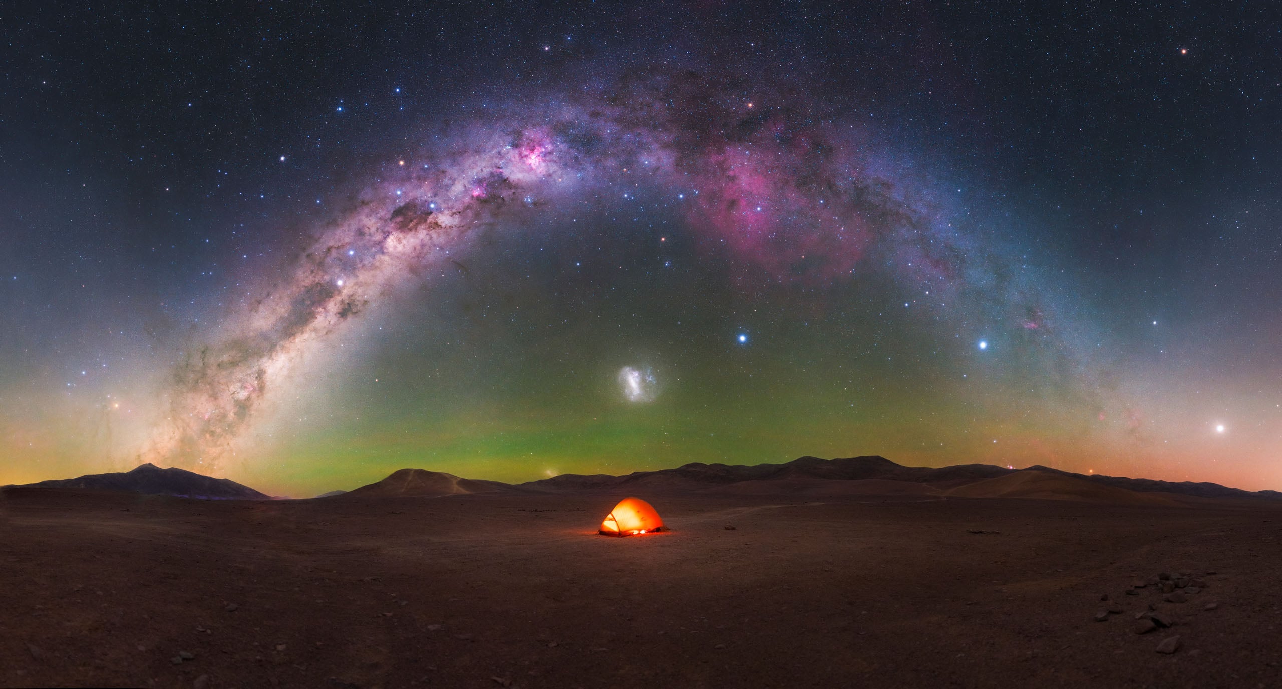 A glowing orange tent sits in a vast, dark desert landscape beneath a panoramic arch of the Milky Way galaxy.