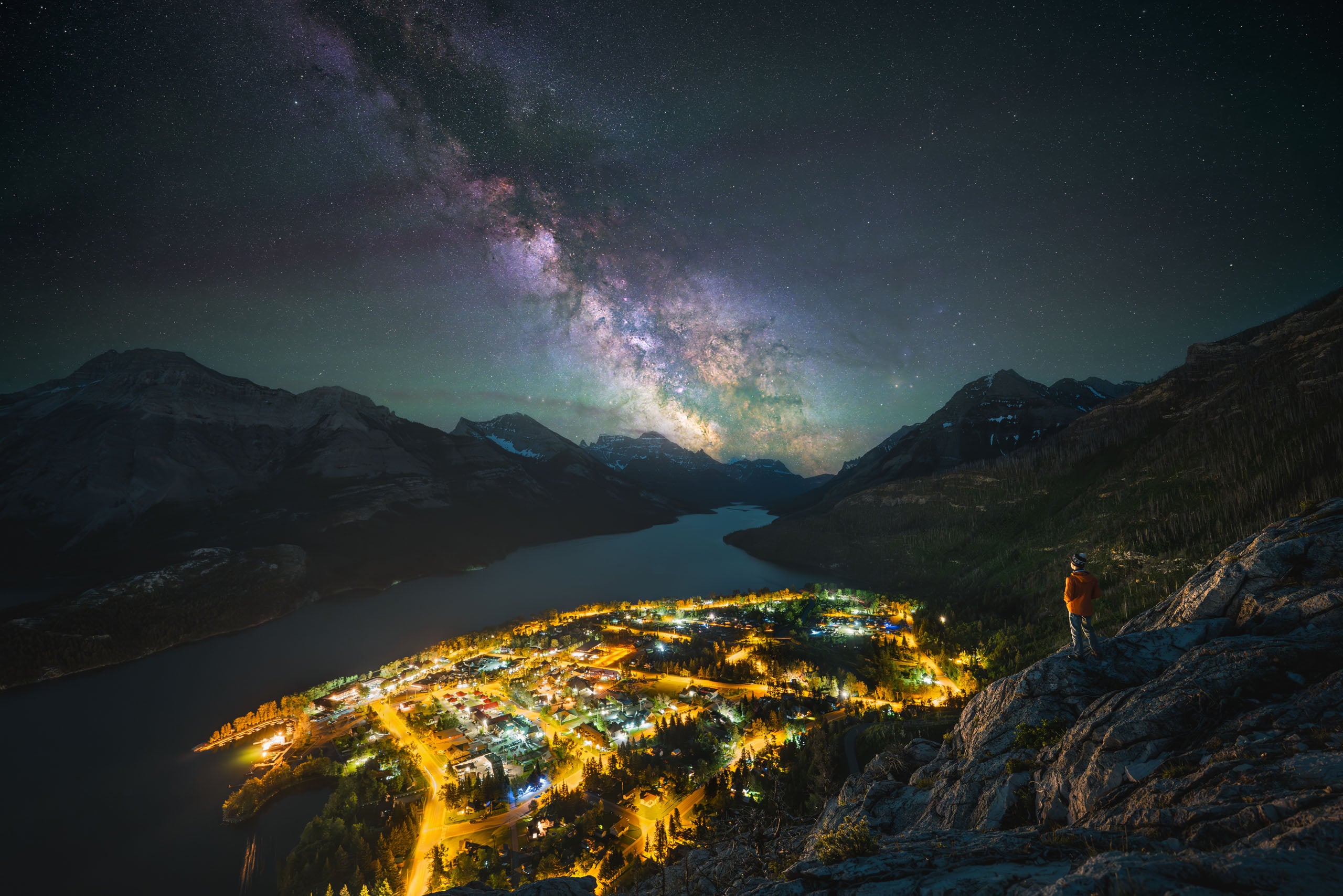 A person in an orange jacket stands on a rocky cliff overlooking a brightly lit town nestled beside a lake under a starry sky.