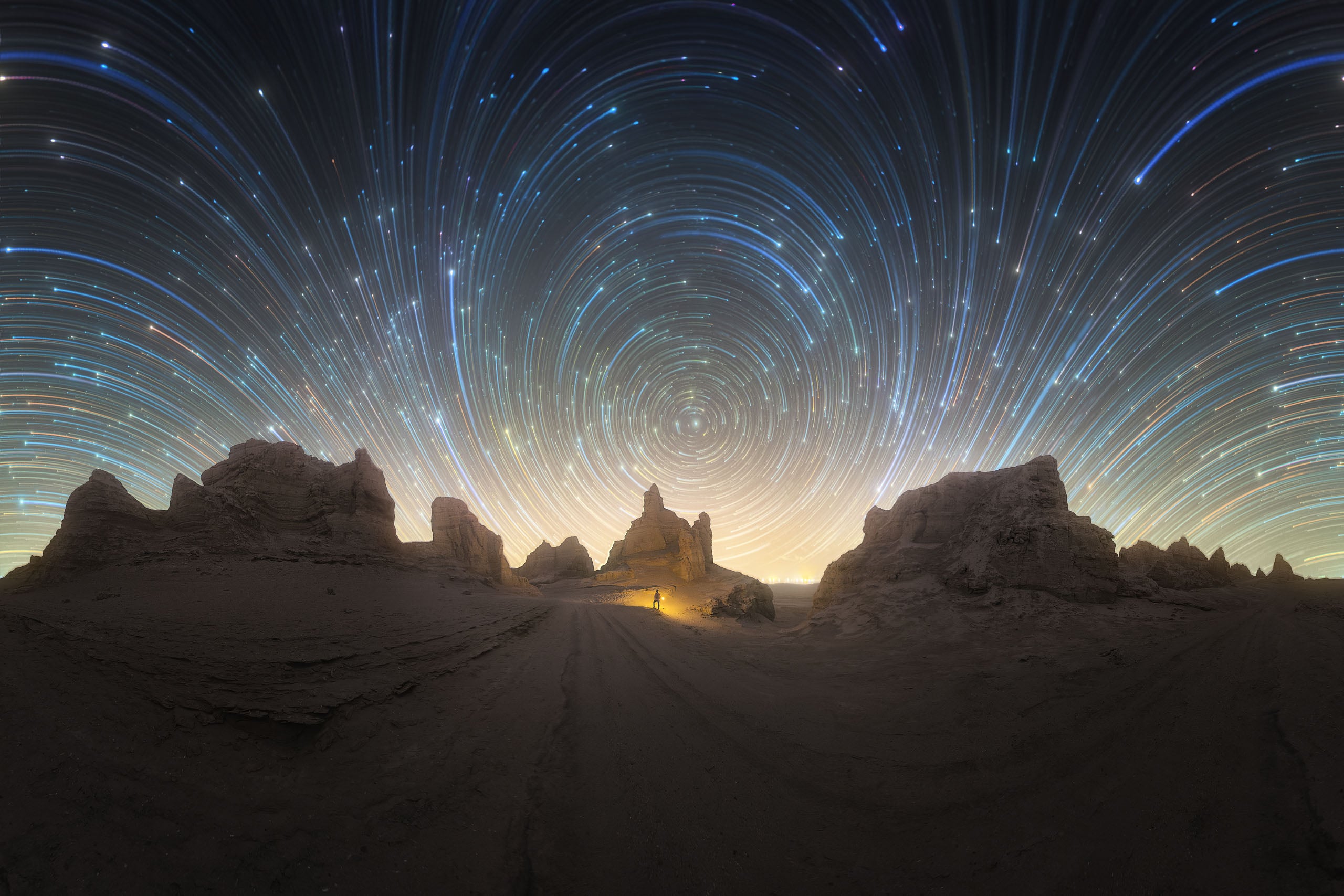 A wide-angle view of a desert landscape at night with a person standing near a rock formation under a sky filled with circular star trails.