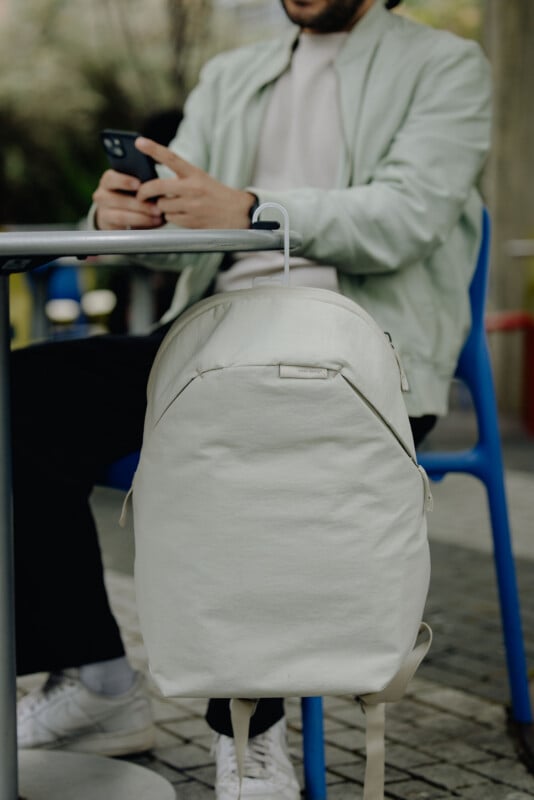 A person sits at a table using a smartphone while a light-colored Peak Design backpack hangs from the edge of the table by a white hook.