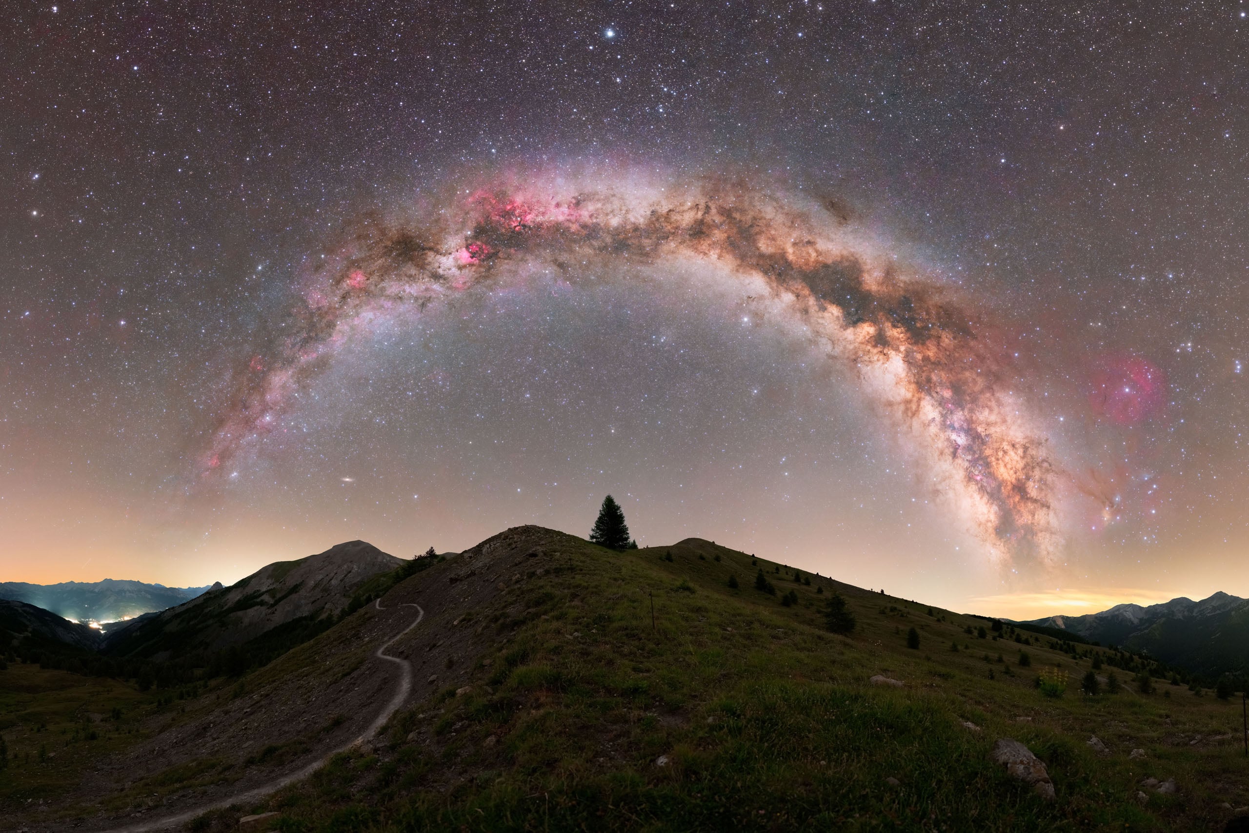 A panoramic view of the Milky Way arching over a grassy mountain ridge with a single tree at the summit.