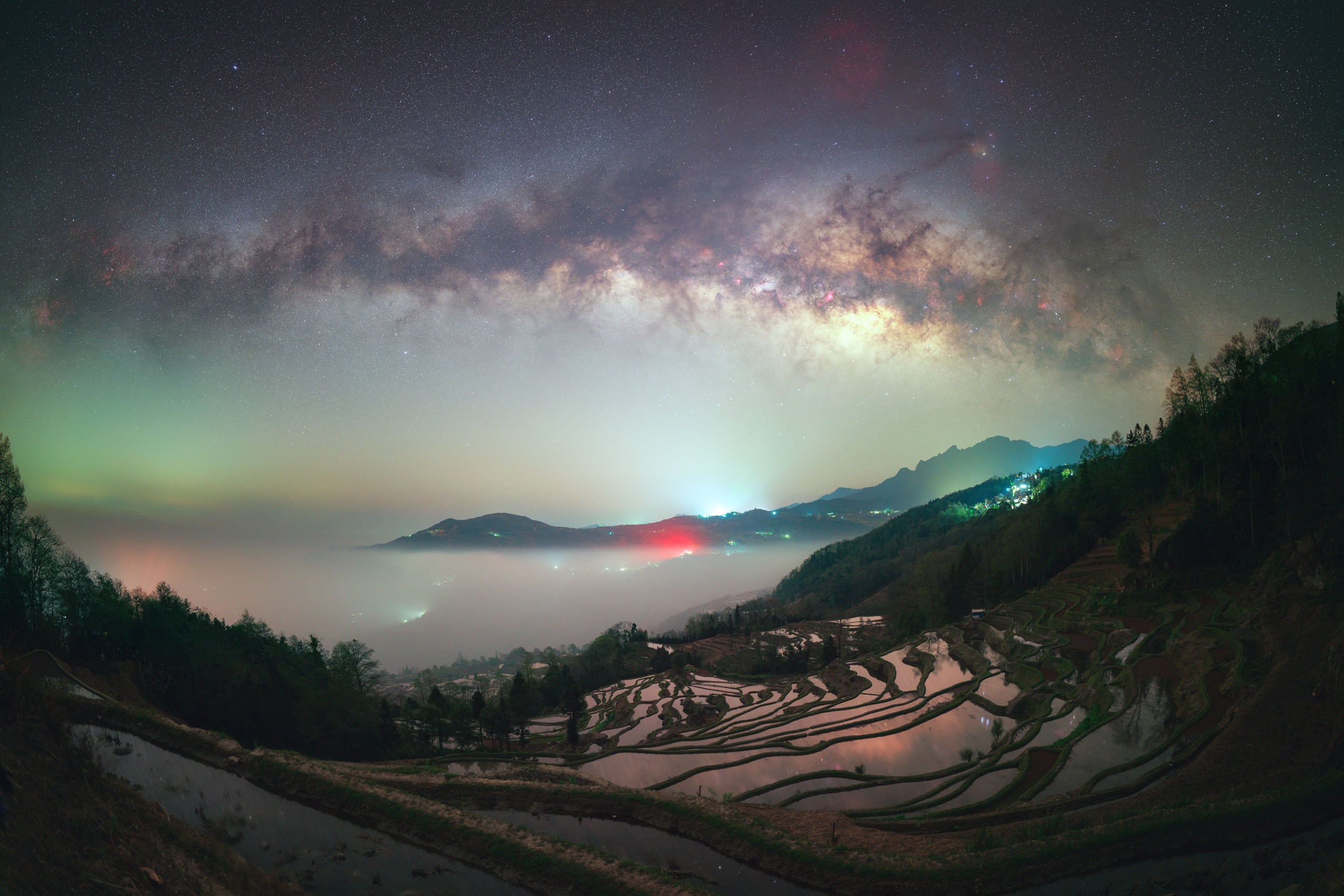 A panoramic view of the Milky Way galaxy arching over terraced rice fields filled with water, with fog settling in the valley.