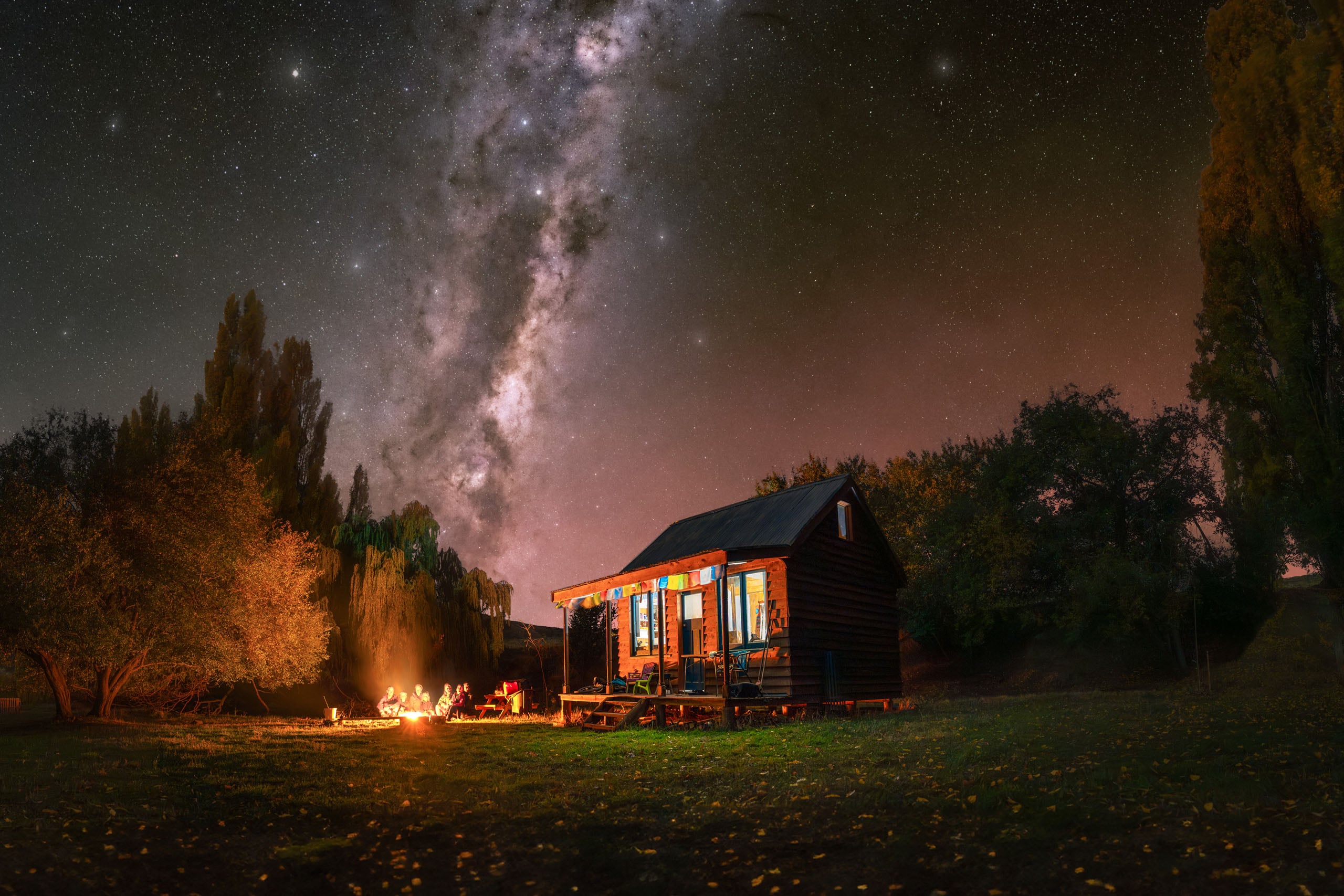 A small wooden cabin sits in a grassy field at night under a bright Milky Way, with a group of people gathered around a campfire.