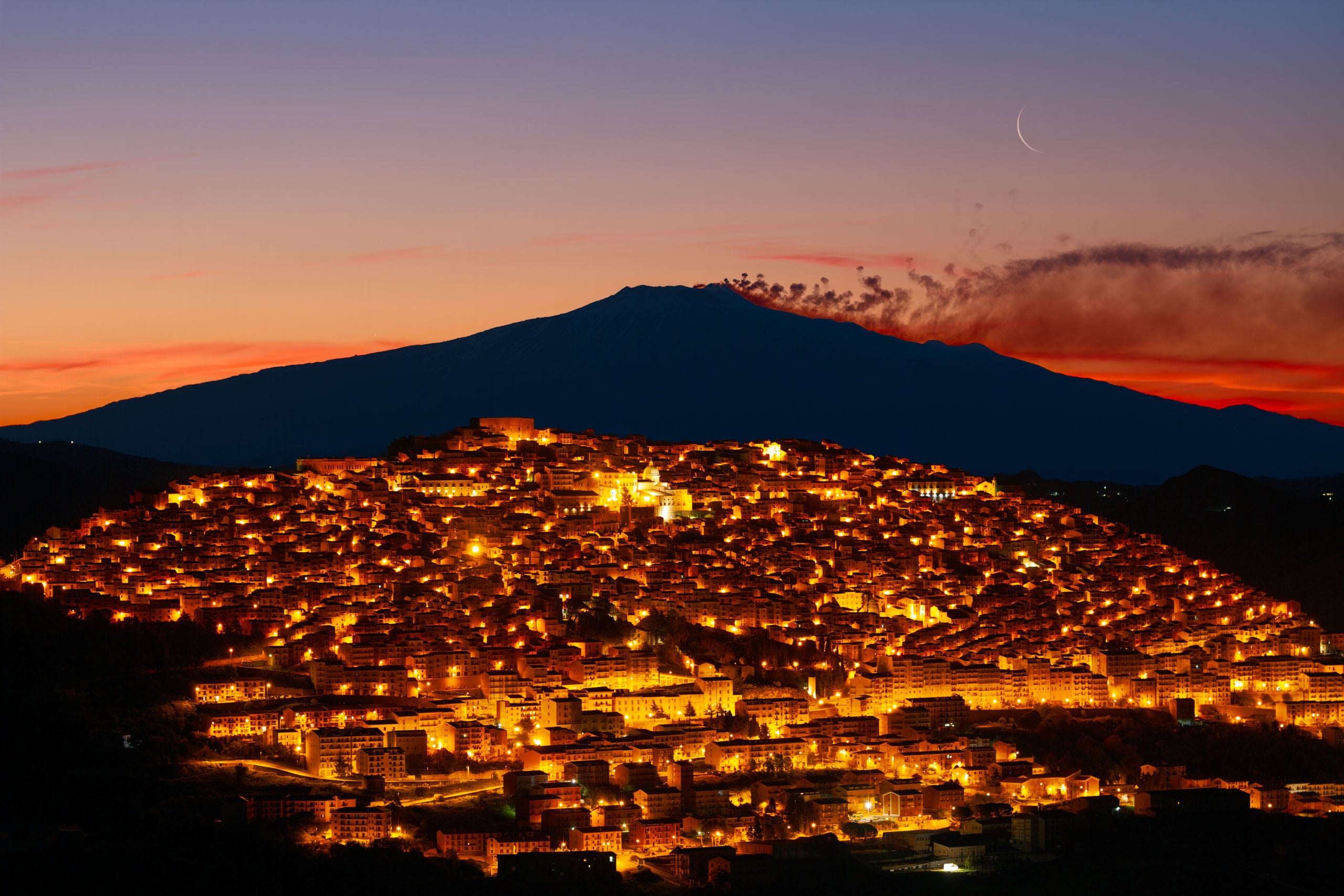 A densely packed town illuminated by warm lights at dusk, set against a large, smoking mountain and a thin crescent moon.
