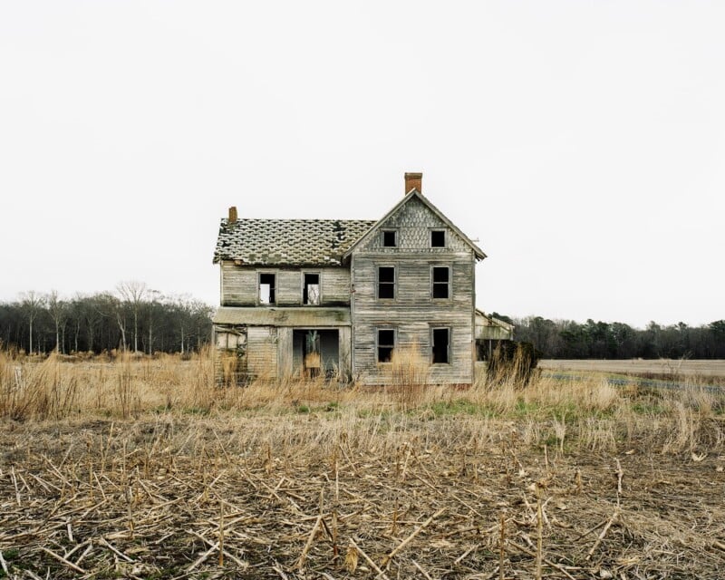 An abandoned, weathered two-story wooden farmhouse stands in the middle of a dry, harvested field under a pale sky.