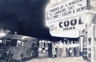 A line of people stands on a sidewalk outside a movie theater at night, with a 