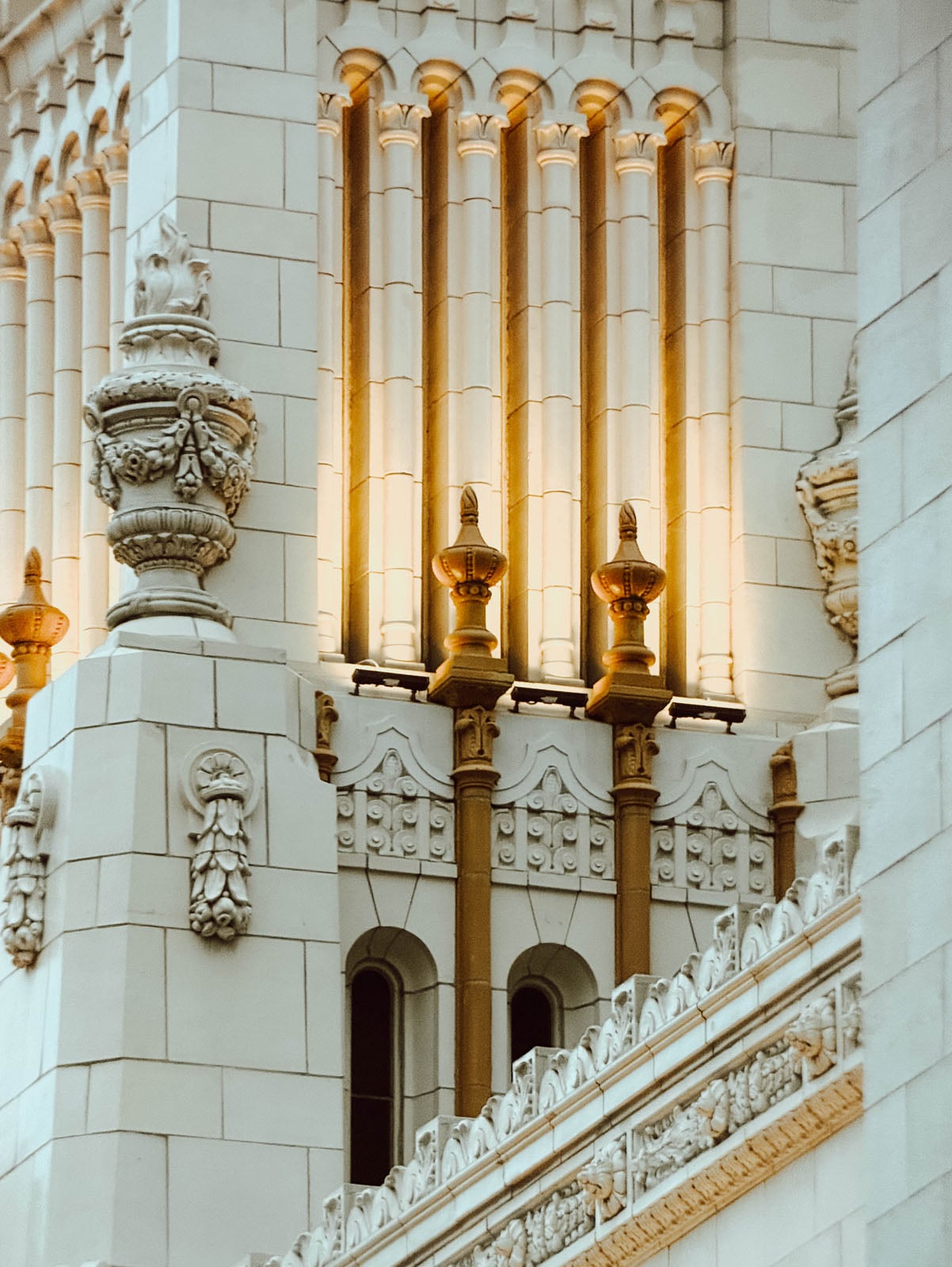 A close-up view of ornate, light-colored stone architecture featuring vertical columns, decorative finials, and intricate carved details illuminated by warm light.