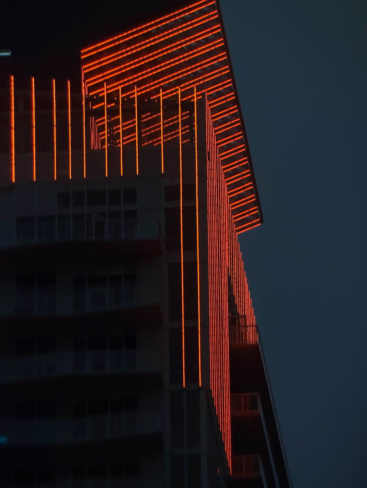 A modern high-rise building at night, illuminated by vertical and horizontal strips of bright orange neon lights along its exterior facade.
