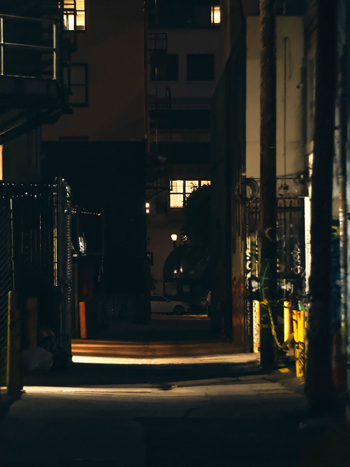 A dimly lit urban alleyway at night, framed by dark buildings and illuminated by a single glowing window and distant streetlights.