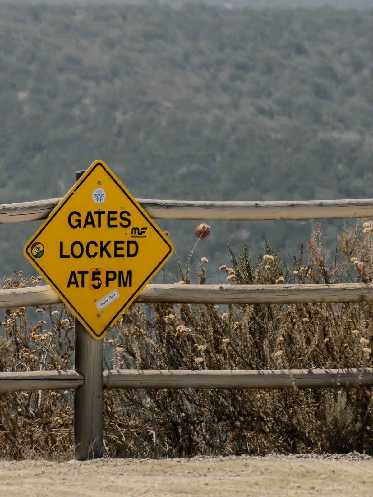 A yellow diamond-shaped sign attached to a wooden fence reads "GATES LOCKED AT 5 PM" with several small stickers applied to its surface.