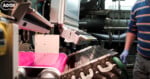 A person wearing blue gloves and a striped sweater operates industrial machinery processing a bright pink material.