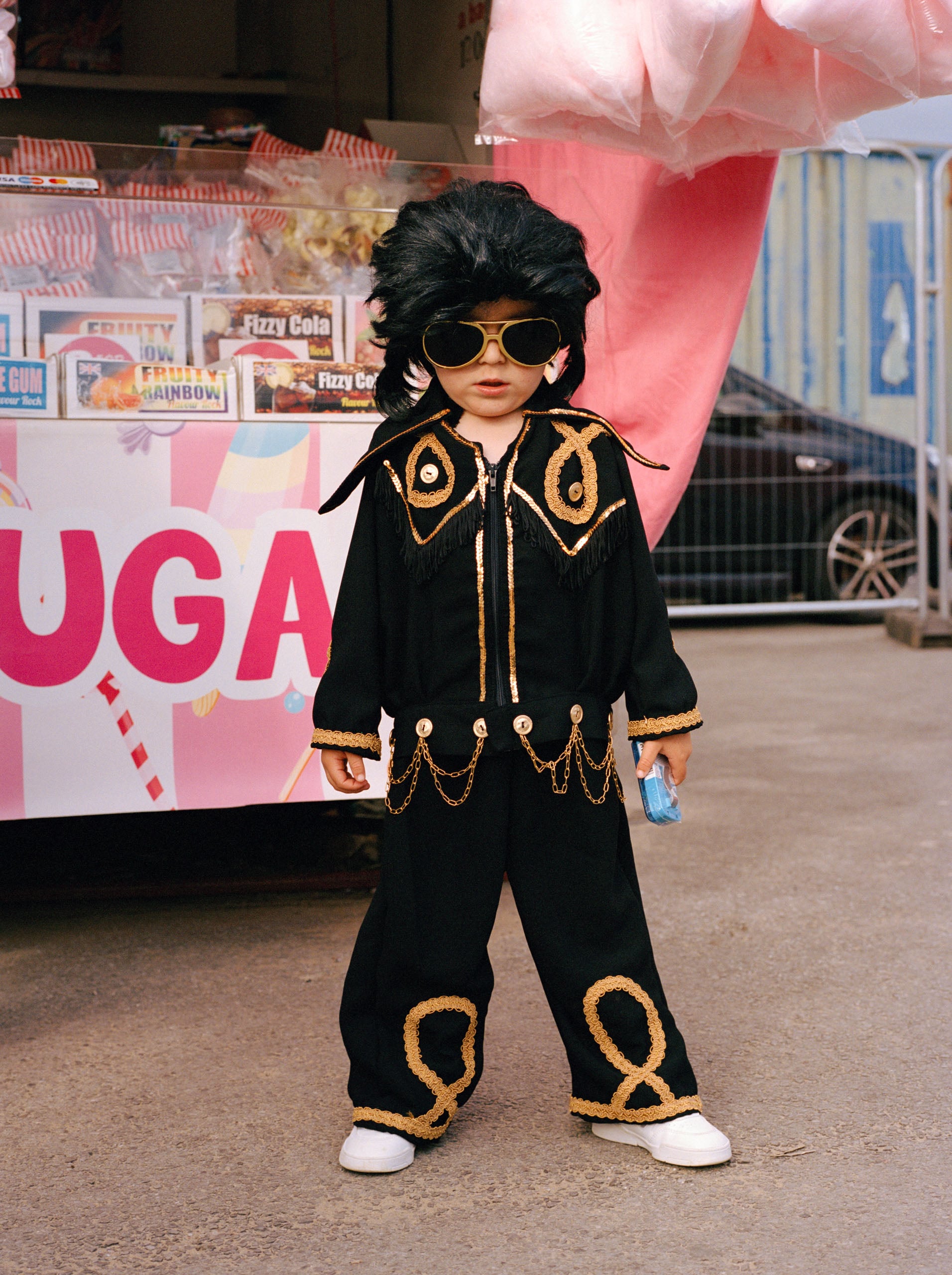 A young child dressed in an Elvis Presley costume, including a black jumpsuit with gold trim, a black wig, and sunglasses, stands in front of a candy stand.