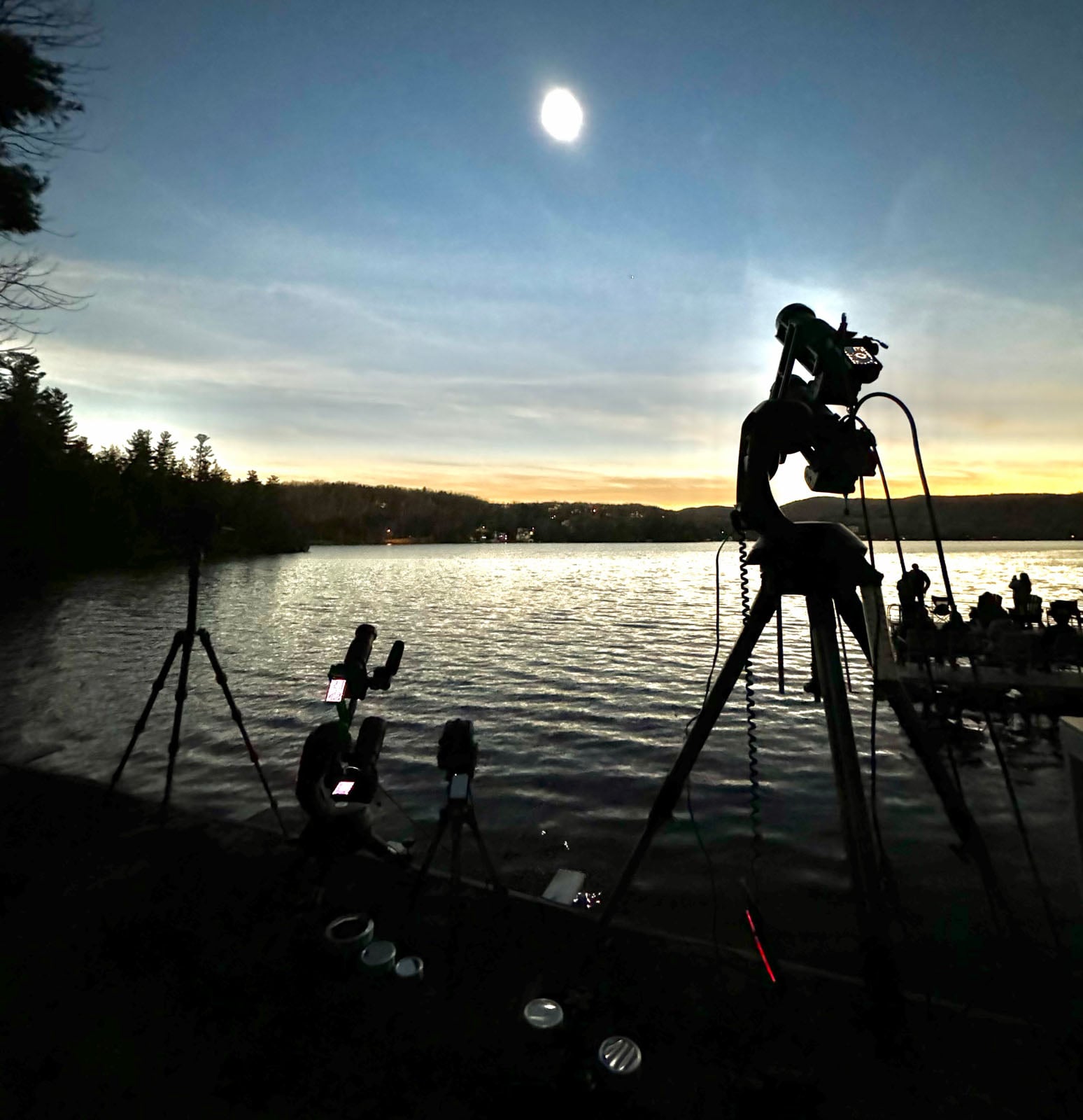 Several tripods with mounted cameras and telescopes are set up along a lakeshore at dusk under a bright moon.
