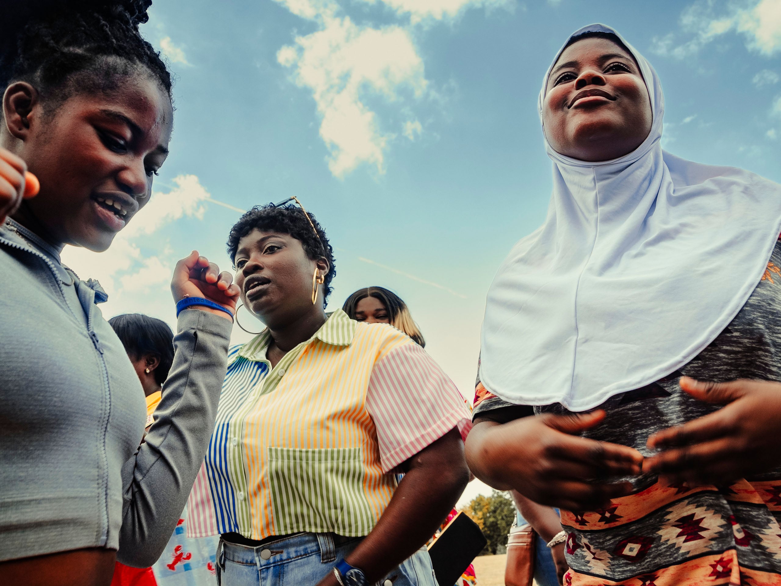 A group of young women stand together outdoors against a blue sky, with one woman wearing a white hijab and another in a colorful striped shirt.