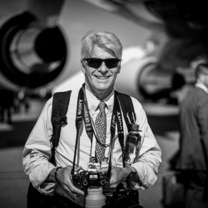 A smiling man with light hair and sunglasses wears a button-down shirt, a patterned tie, and a camera strap around his neck while holding a camera in front of a large aircraft engine.