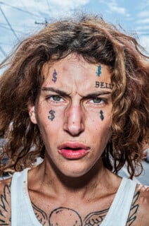 A close-up portrait of a person with curly brown hair, facial tattoos including dollar signs and the word