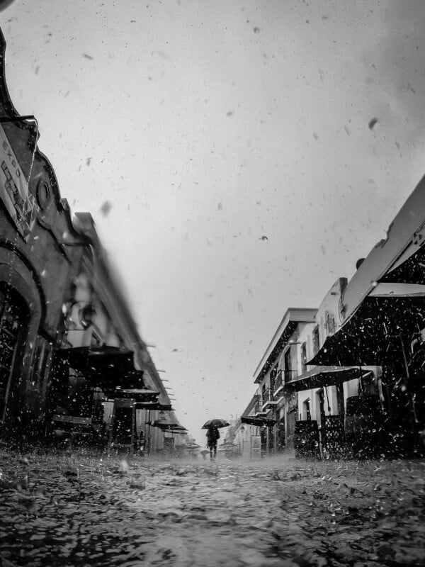 A person with an umbrella walks down a rain-slicked street between buildings during a heavy downpour.