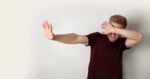 A man with reddish-brown hair and a beard wearing a maroon t-shirt holds one hand out in a stop gesture while covering his eyes with his other hand.