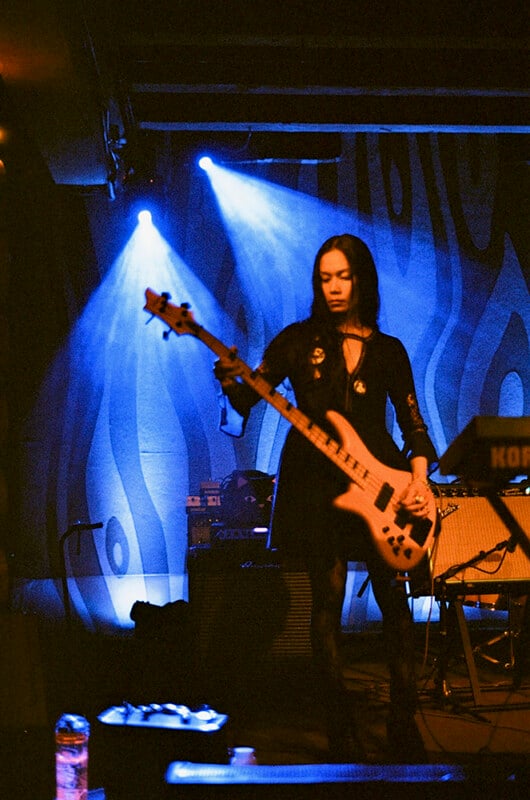 A person with long dark hair plays a light-colored bass guitar on a stage illuminated by blue spotlights against a patterned backdrop.