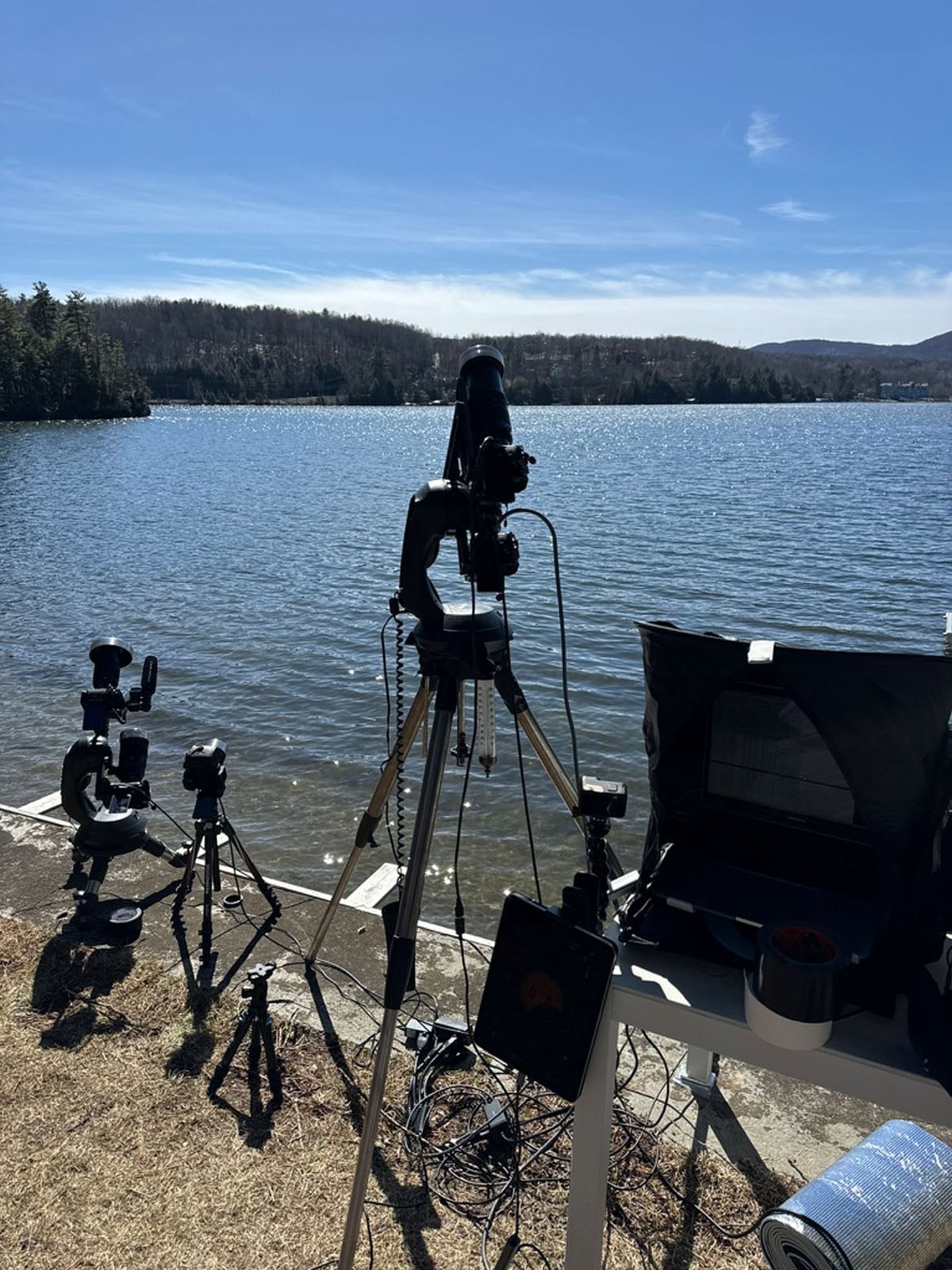 Telescopes and camera equipment are set up on tripods along a lakeshore on a sunny day, with a laptop under a shade hood.