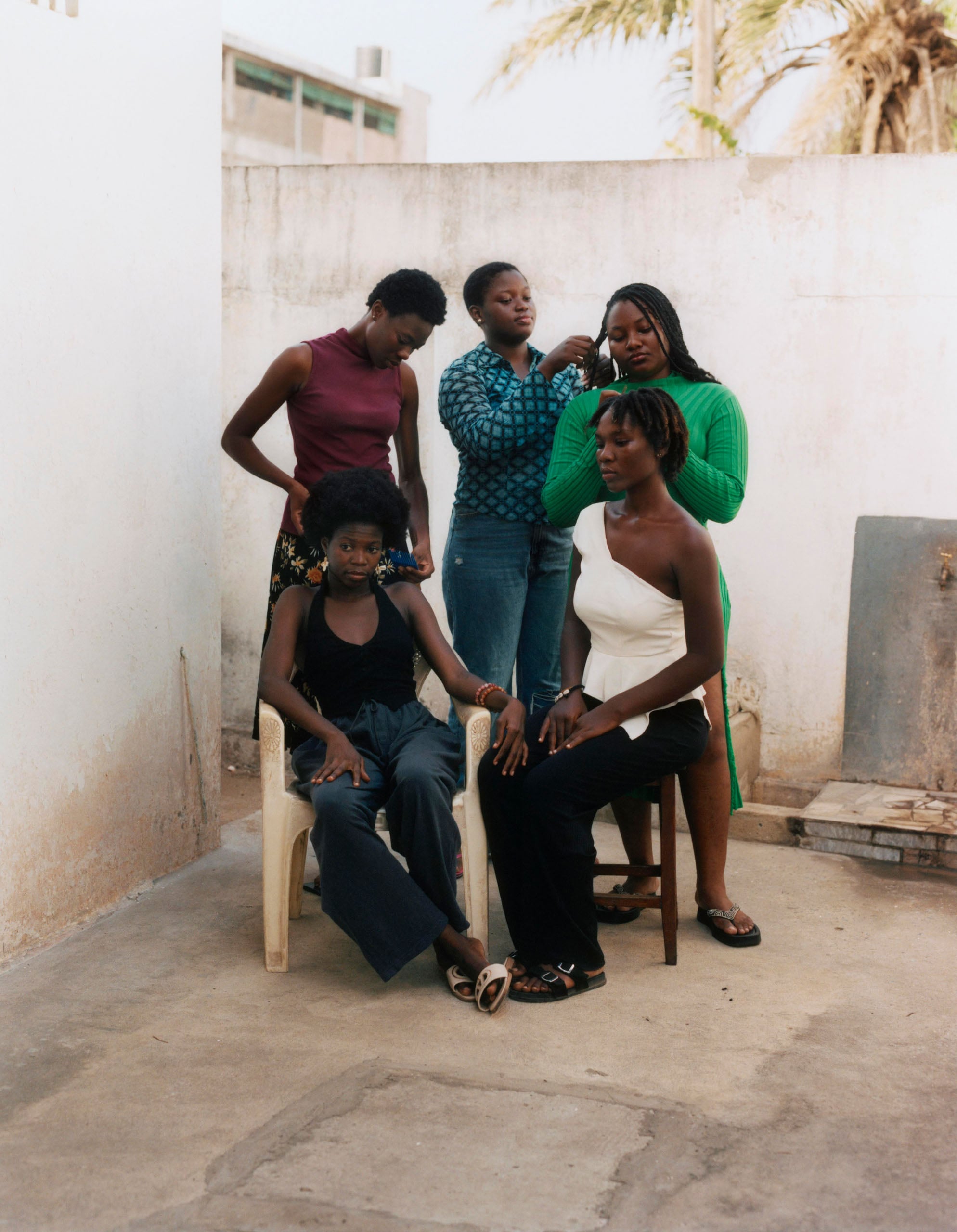 Four young Black women gather outdoors, with two seated in chairs while the others stand behind them styling their hair.