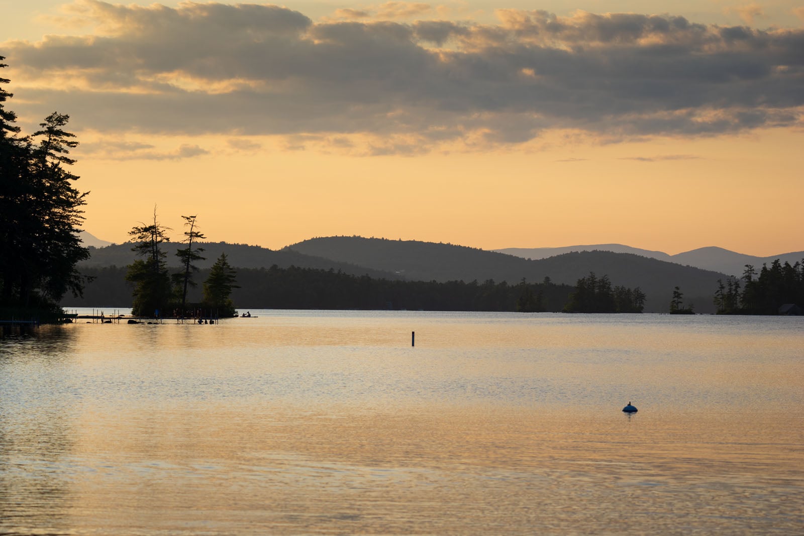 A calm lake at sunset with silhouetted trees on the shore, distant mountains, and a soft orange sky with scattered clouds.