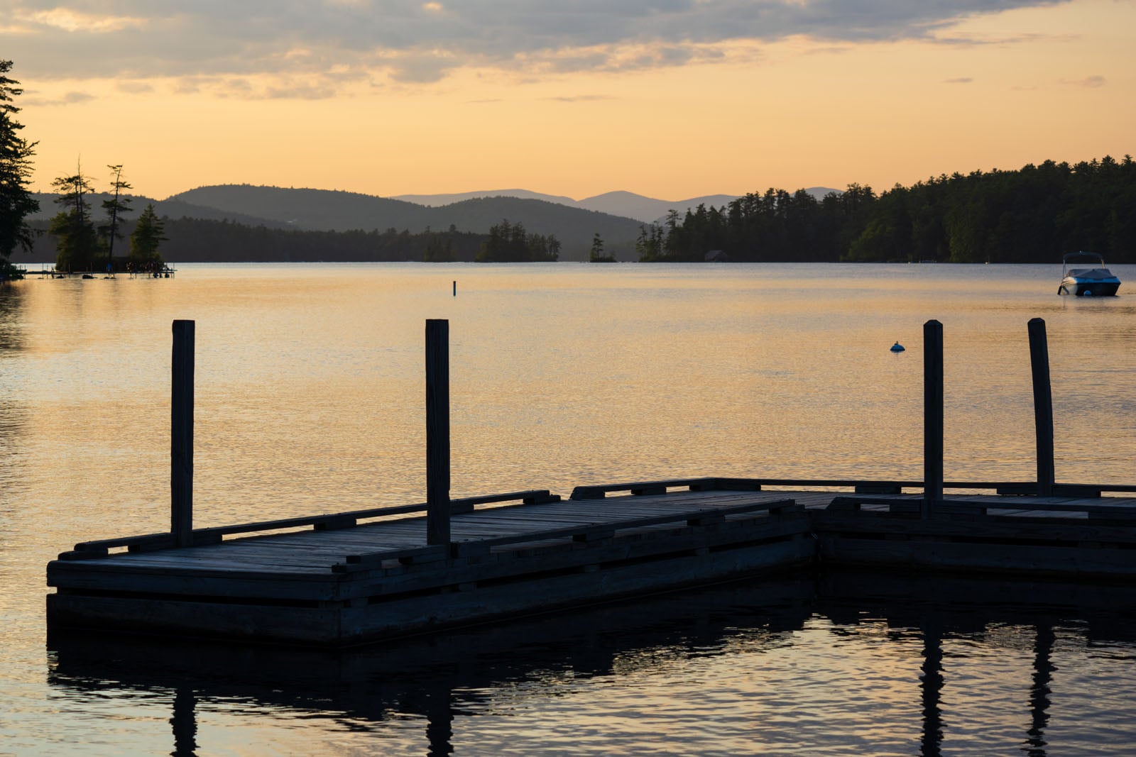 A wooden dock extends into a calm lake at sunset, with distant mountains and a small boat visible on the water.