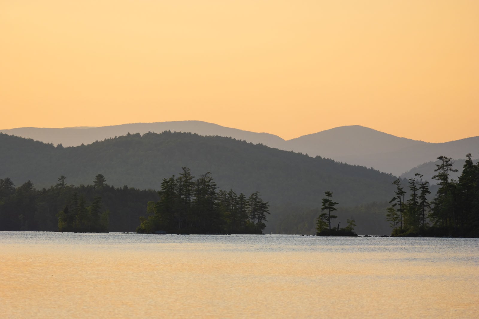 A calm lake reflects a golden sunset, with silhouettes of forested islands and distant mountain ranges in the background.