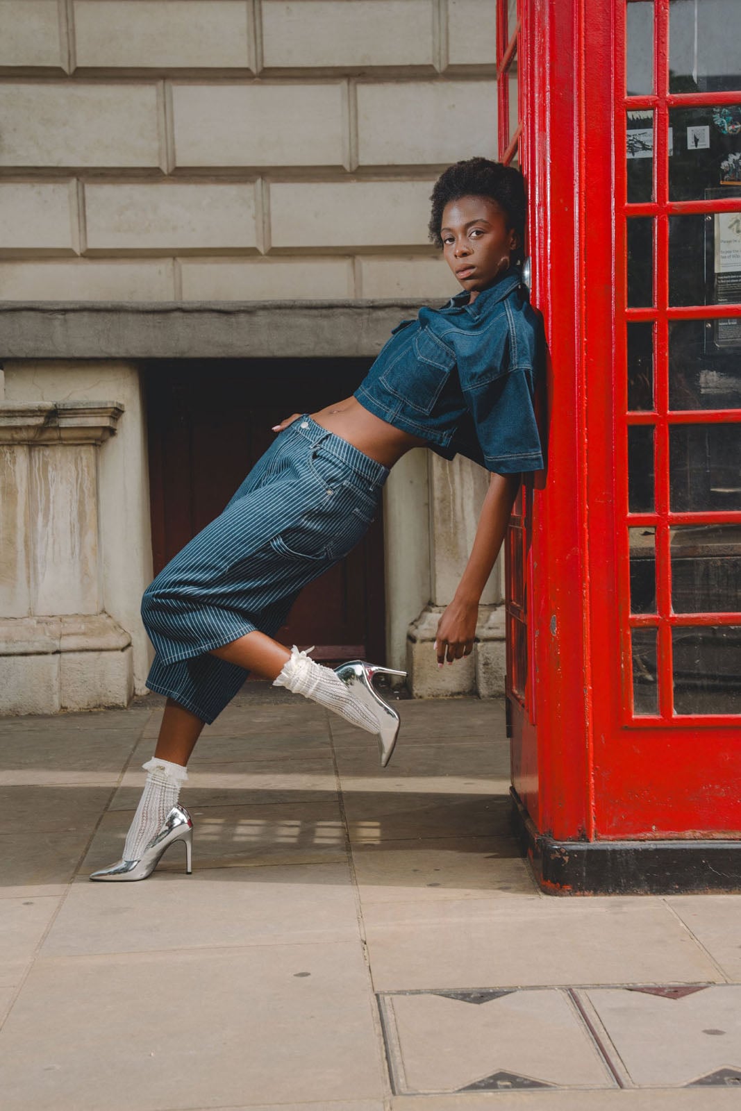 A person with short dark hair leans against a red telephone booth, wearing a cropped denim jacket, pinstriped culottes, white ruffled socks, and silver high heels.