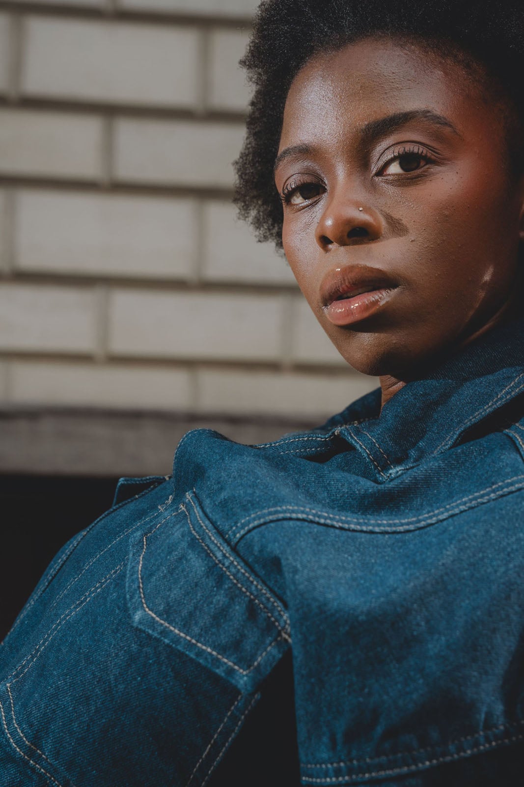 A close-up portrait of a Black woman with short, dark hair wearing a denim jacket, looking directly at the camera.