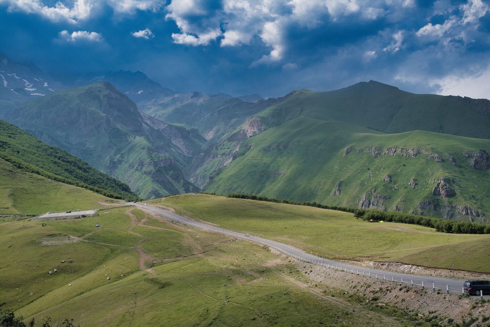 A paved road winds through a vast, green mountain valley under a dramatic sky with dark, heavy clouds.