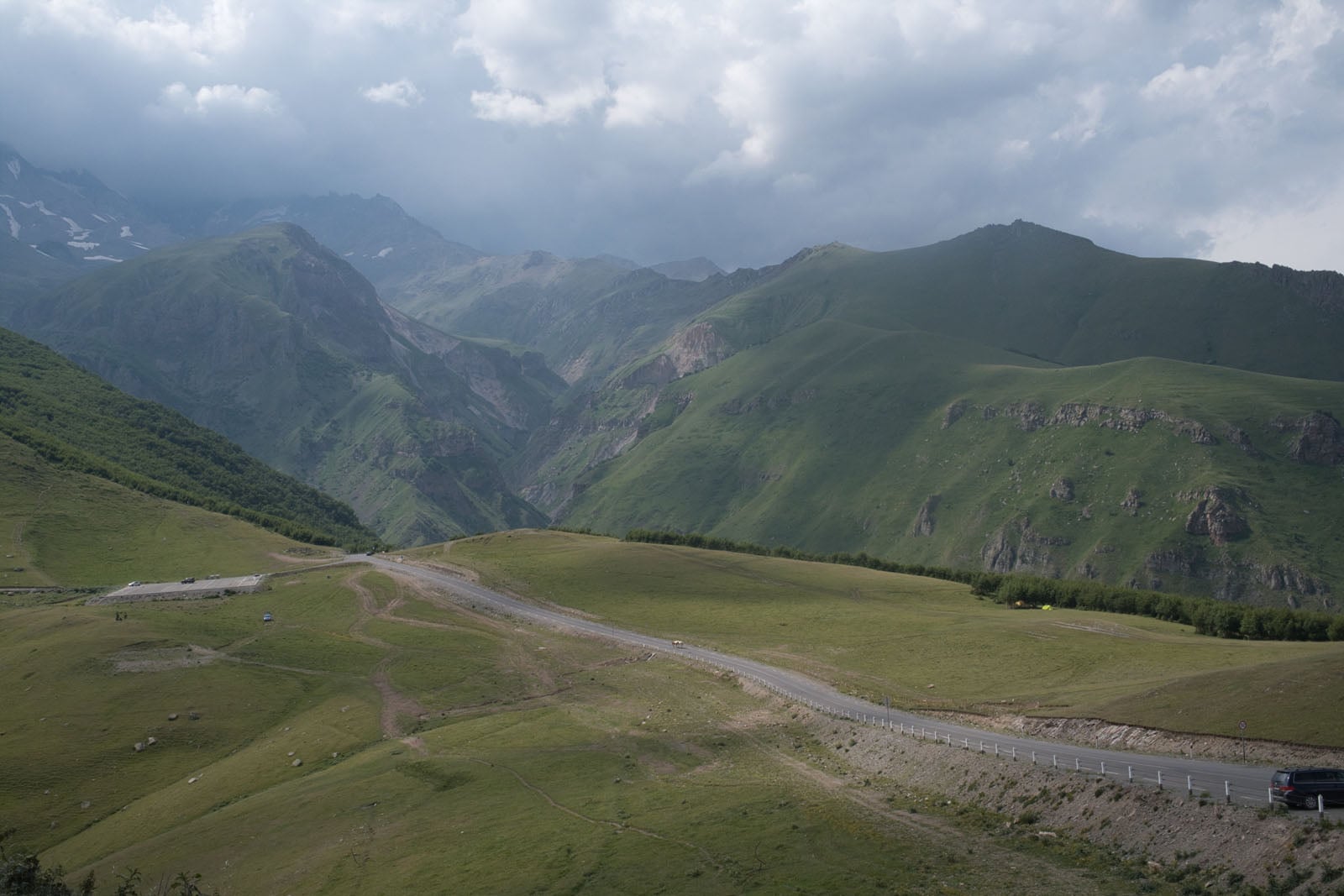 A paved road winds through a vast, grassy mountain landscape under a cloudy sky, with a dark SUV parked on the right shoulder.