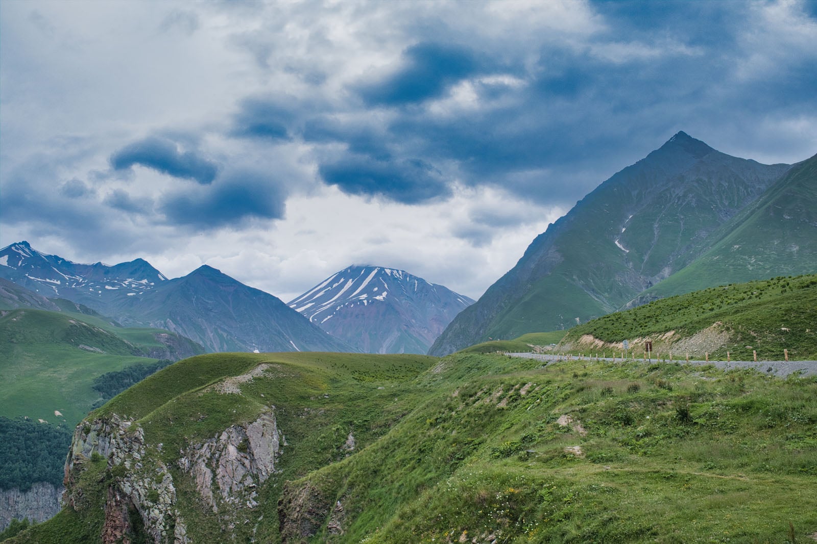 A winding road cuts through a lush green mountain landscape under a cloudy, dramatic sky with snow-capped peaks in the distance.