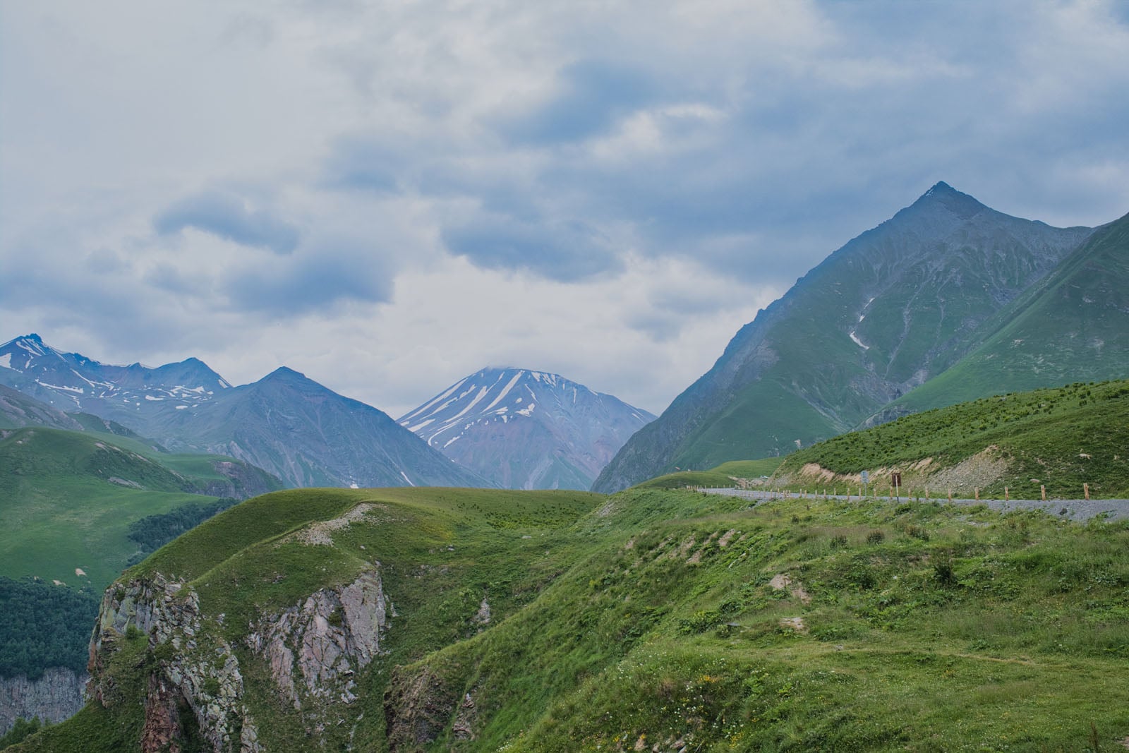A winding road traverses a lush green mountain landscape under a cloudy sky, with snow-capped peaks visible in the distance.