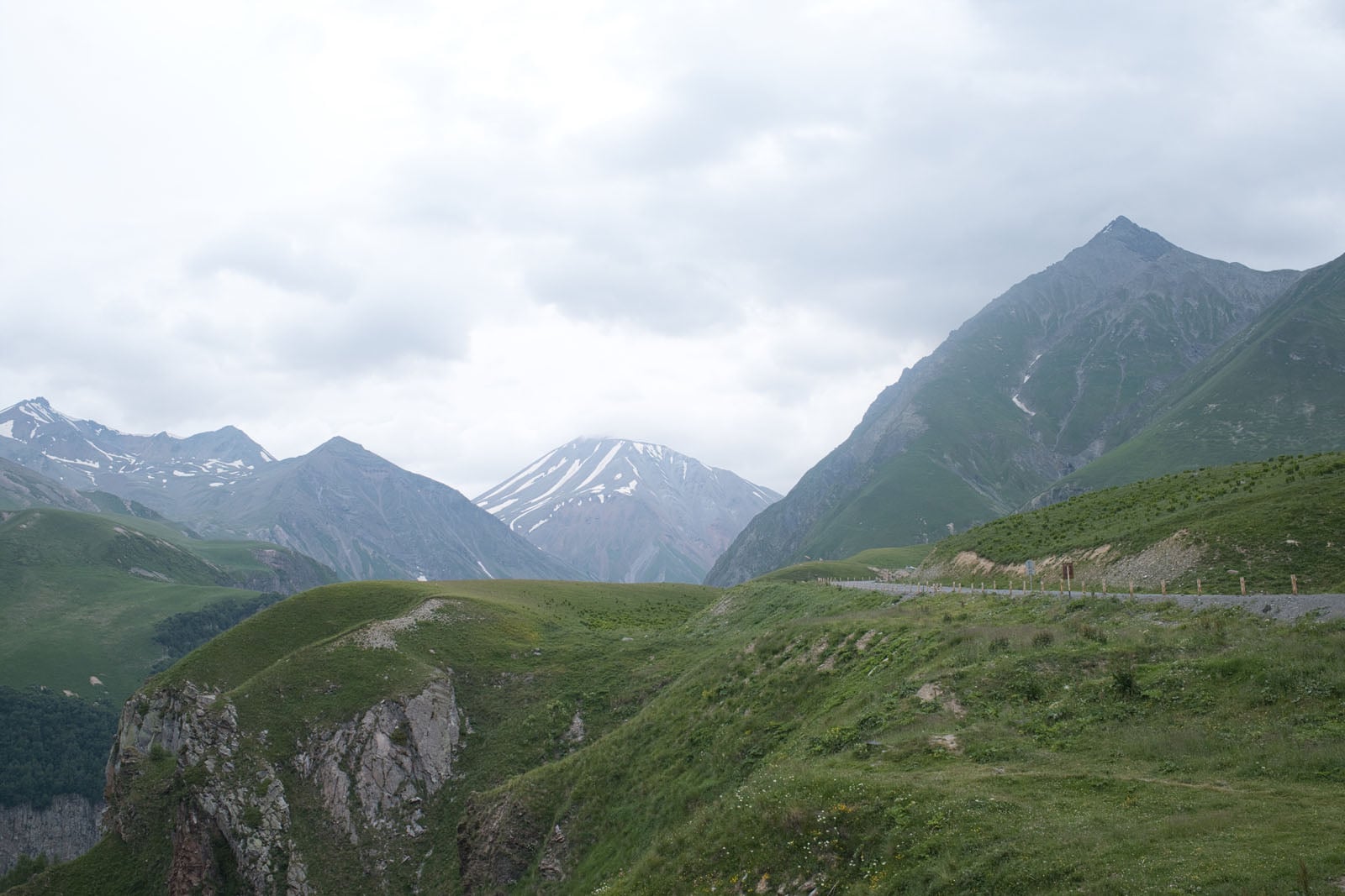 A winding road traverses a green, mountainous landscape under a cloudy sky, with snow-capped peaks in the distance.