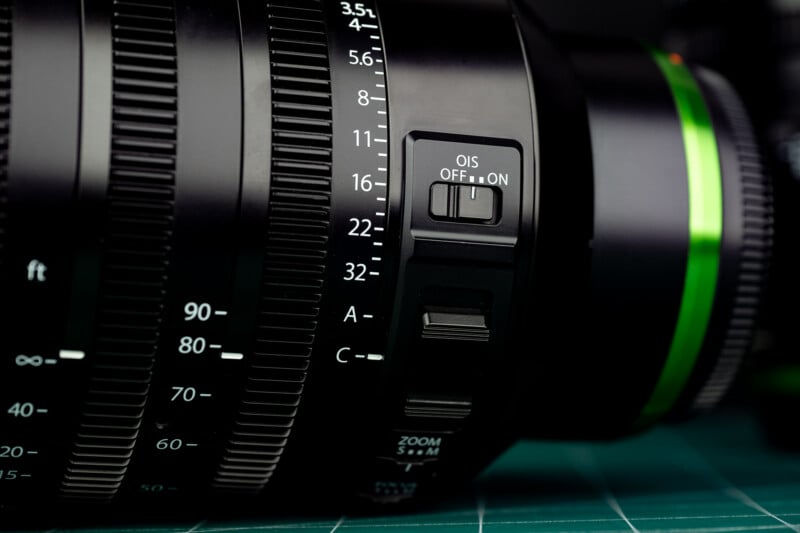 A close-up view of a camera lens barrel showing aperture settings, a zoom ring, and an OIS switch set to ON.