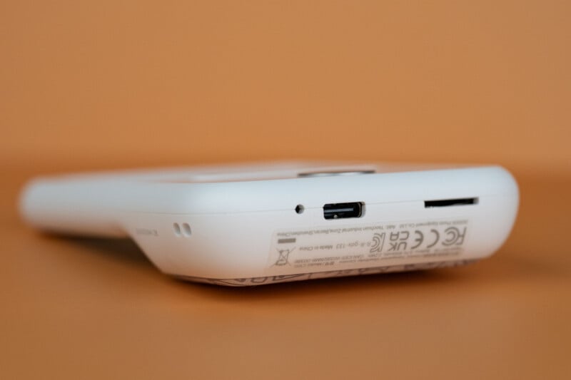A white digital camera rests on an orange surface, showing its bottom edge with a USB-C charging port, a memory card slot, and regulatory text.