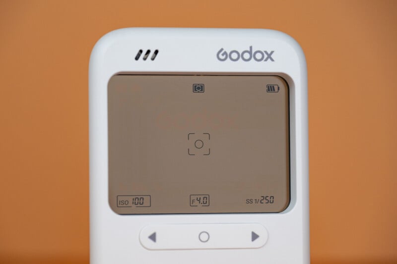 A close-up of a white Godox device with a digital screen displaying camera settings: ISO 100, F4.0, and SS 1/250.
