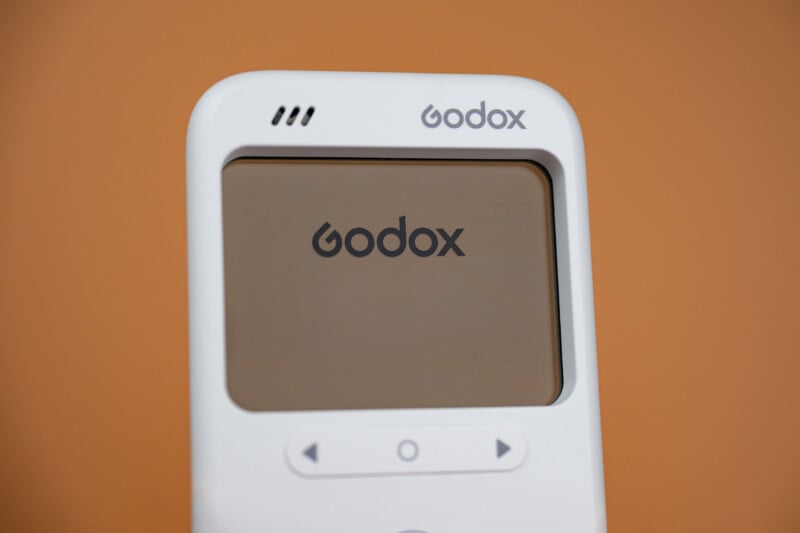 A white Godox device with a screen displaying the Godox logo, set against a solid orange background.