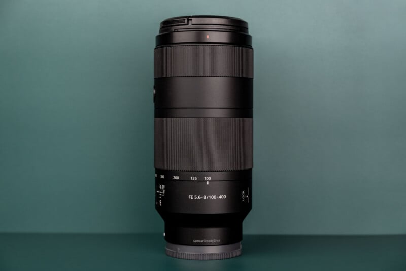 A black telephoto zoom lens stands vertically against a solid teal background, with the text "FE 5.6-8/100-400" visible on its barrel.