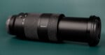A black Sony telephoto zoom lens with a lens cap attached, resting horizontally on a dark green surface.