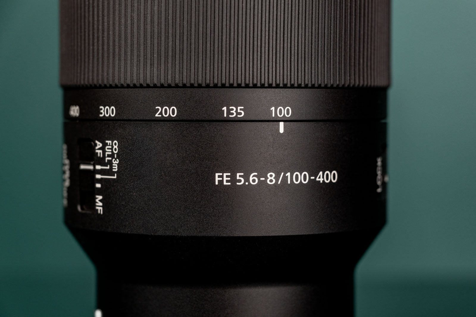 A close-up of a camera lens barrel showing a zoom ring set to 100mm, with focus and aperture markings visible.