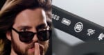 A man with long hair and a beard holds a finger to his lips while wearing sunglasses, shown next to a close-up of a device arm displaying battery, camera, Wi-Fi, and notification icons.