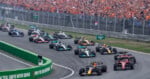 A pack of Formula 1 race cars navigates a turn on a track in front of a large crowd of spectators.